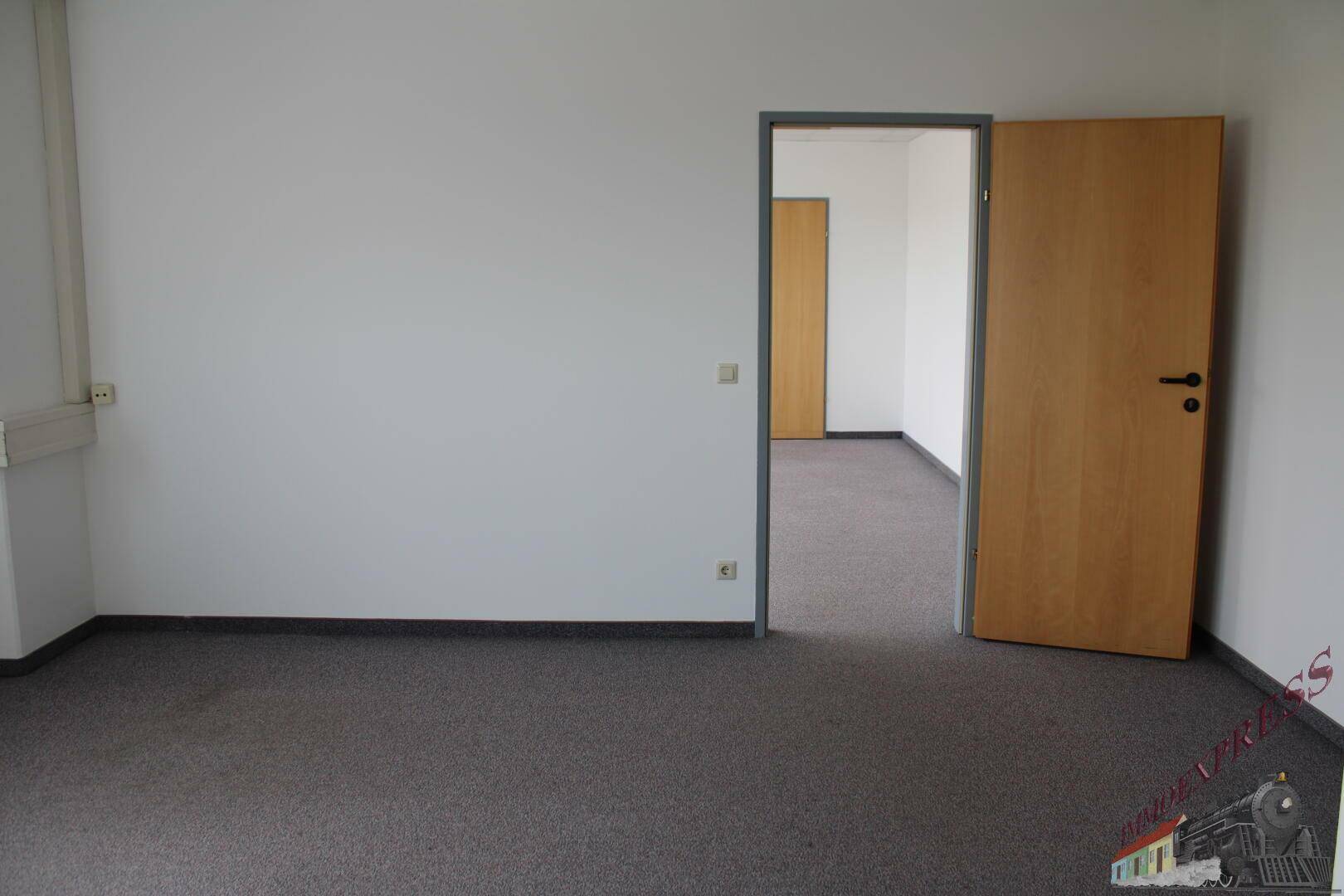 Leerer Büroraum mit Teppichboden und einer offenen Tür, die den Blick in einen weiteren Raum freigibt.