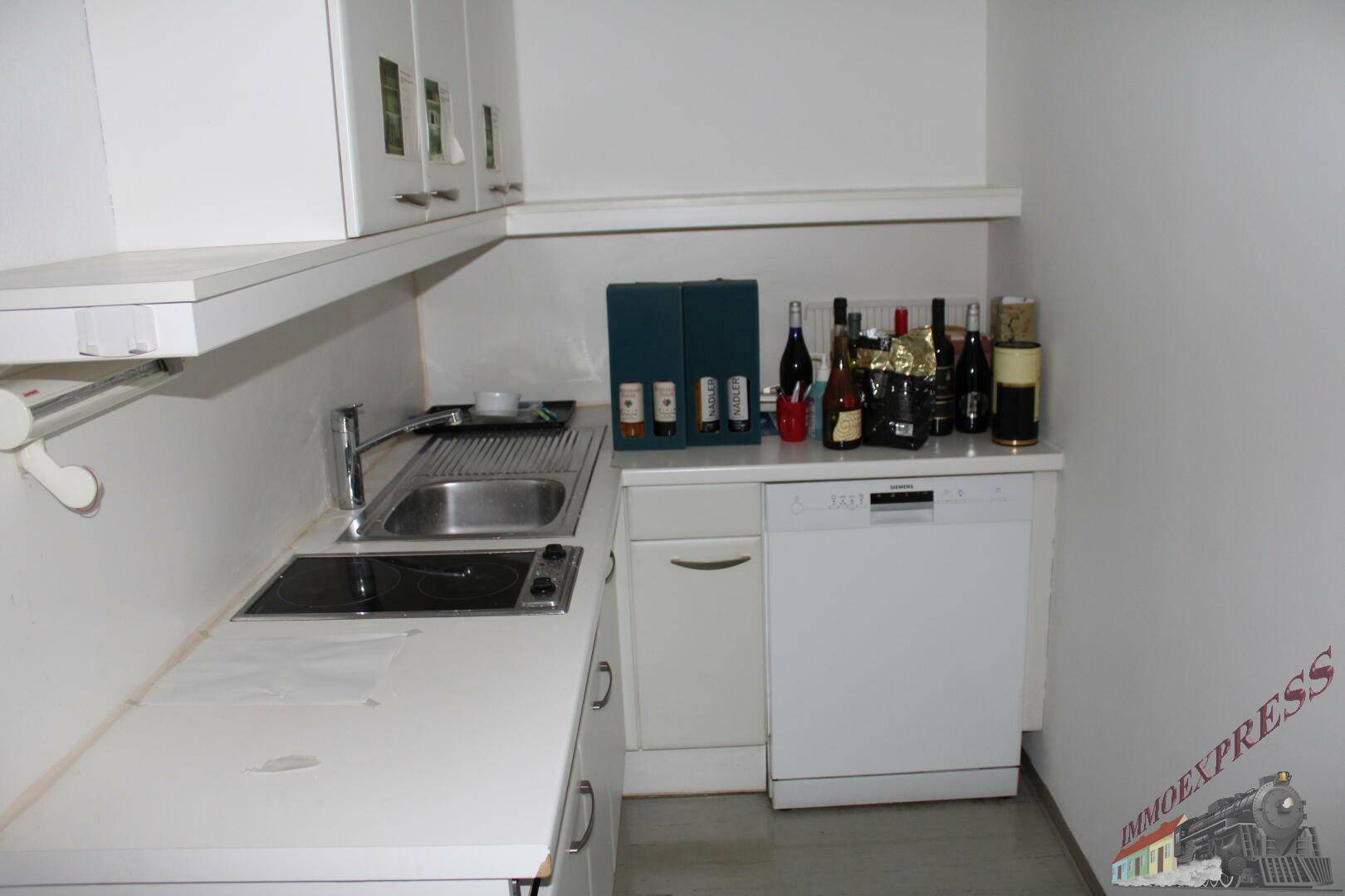 Kleine Küchenzeile mit Spüle, Geschirrspüler und weißen Hängeschränken, ideal für eine Büroküche.