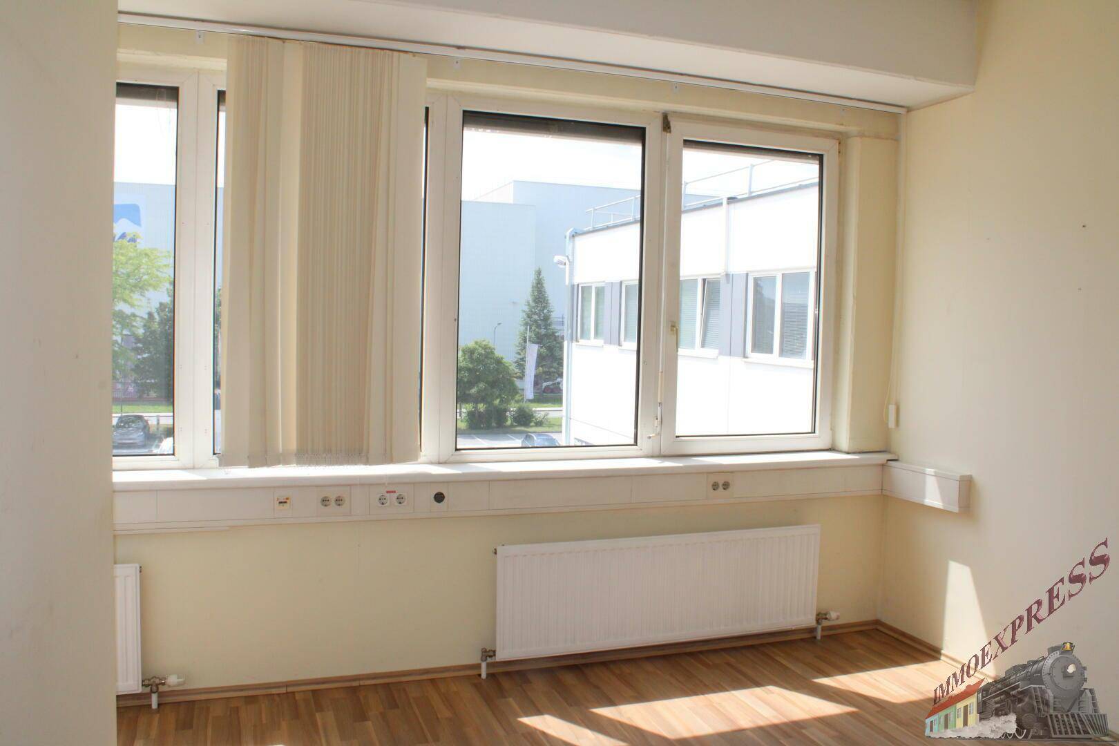 Helles Büro mit Holzboden und großen Fenstern, die einen Blick auf das umliegende Gebäude bieten.