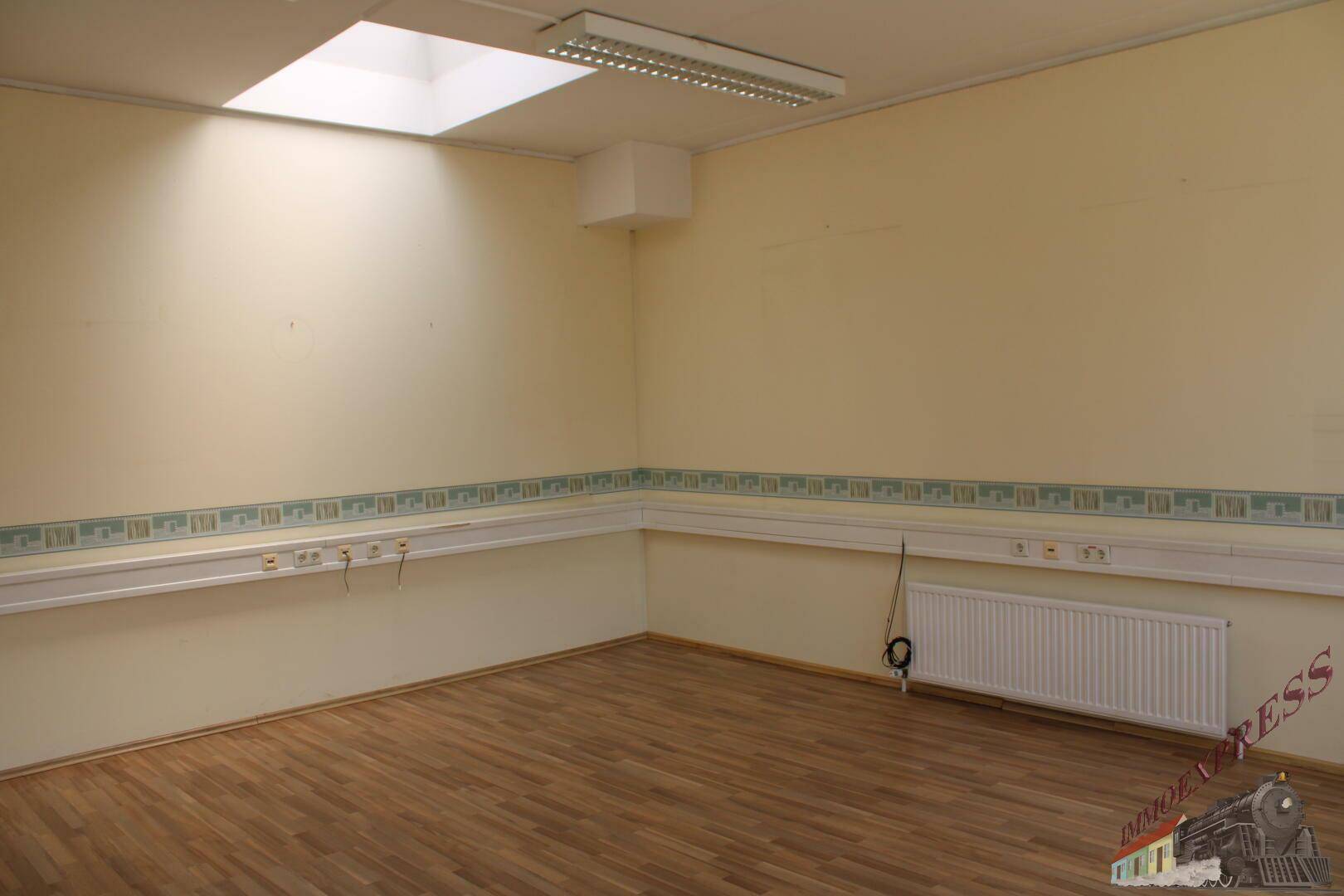 Leeres Büro mit Holzfußboden, Deckenfenster und einer umlaufenden Steckdosenleiste.