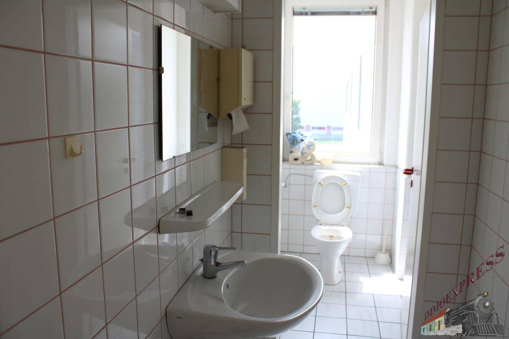 Helles Badezimmer mit weißen Fliesen, Waschbecken und Toilette, ausgestattet mit einem Fenster.