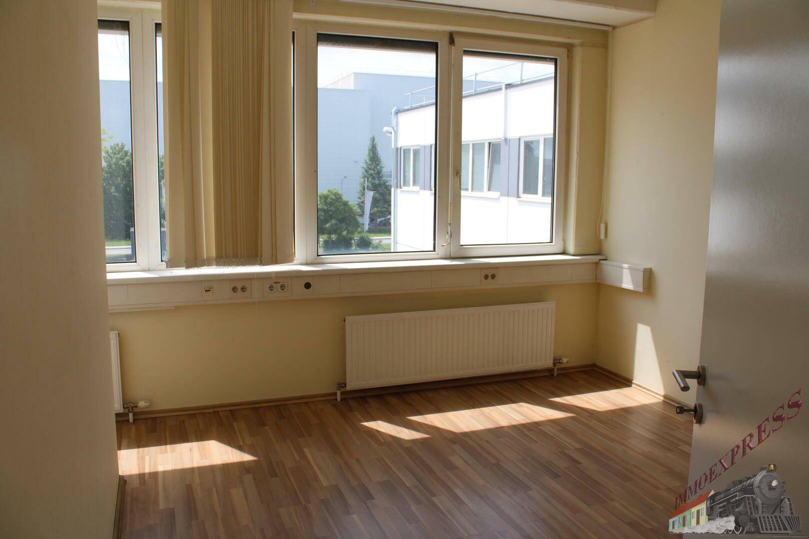 Heller Büroraum mit großen Fenstern, Holzfußboden und Heizkörper, bietet viel Tageslicht.