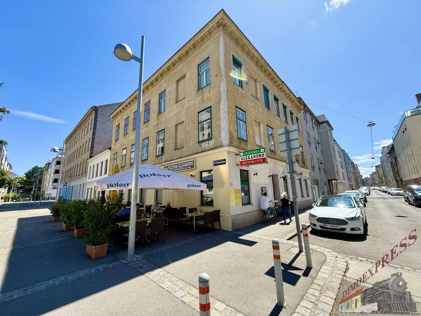 Weitwinkelaufnahme eines Eckgebäudes mit einer Pizzeria und ihrer Außenterrasse, umgeben von städtischer Umgebung.