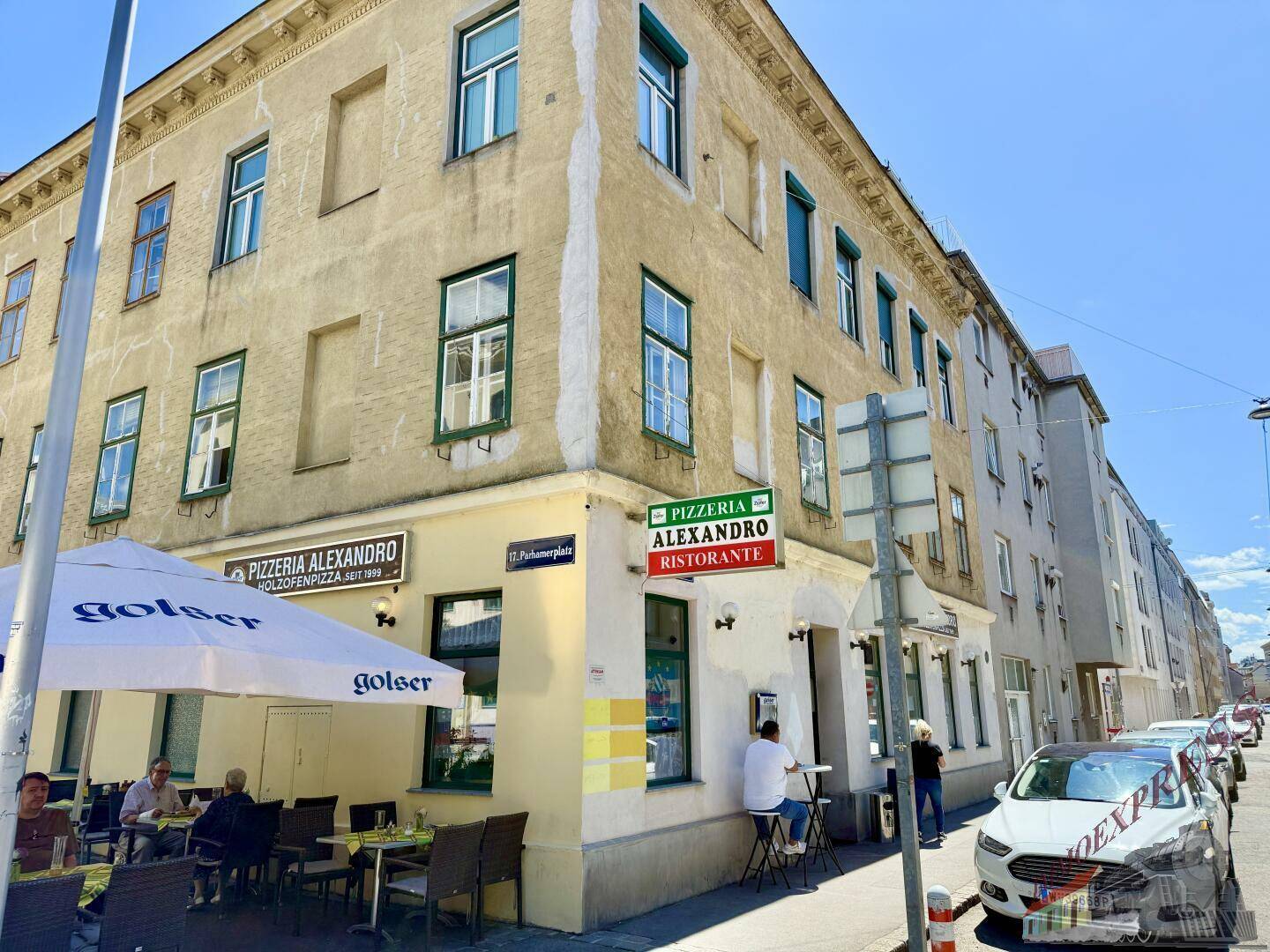Weitwinkelansicht eines Eckgebäudes mit einer Pizzeria und ihrer Außenterrasse, umgeben von städtischer Architektur.