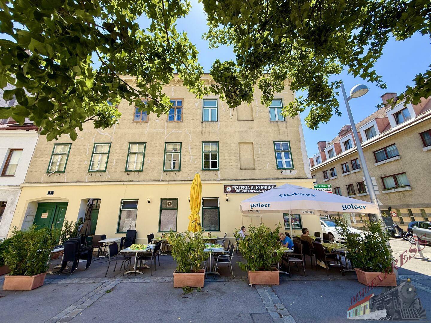 Frontansicht eines Gebäudes mit einer Pizzeria im Erdgeschoss und einer Außenterrasse, teilweise von Bäumen verdeckt.
