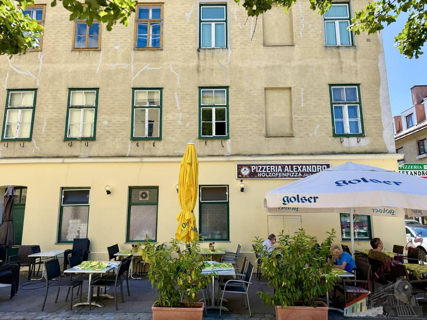 Fassadenansicht eines Gebäudes mit einer Pizzeria im Erdgeschoss und einer Außenterrasse, beschattet von Bäumen.