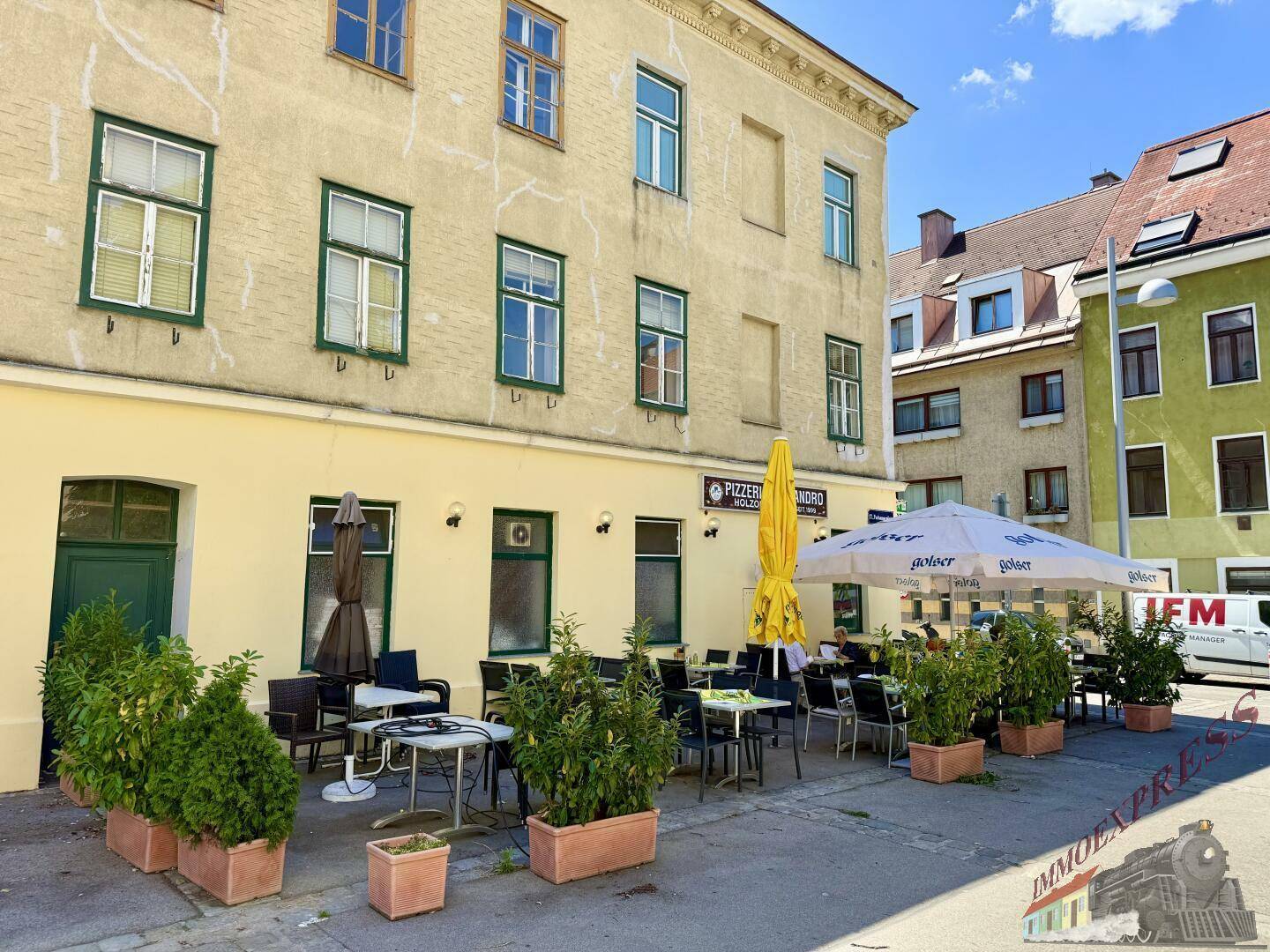 Blick auf die Fassade eines Gebäudes mit einer Pizzeria im Erdgeschoss und einer möblierten Außenterrasse mit Pflanzen.