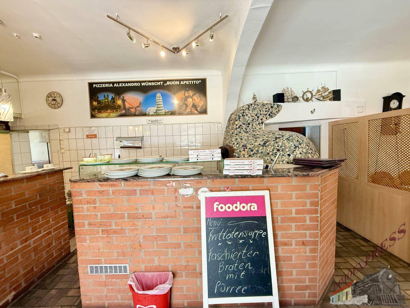Servicebereich einer Pizzeria mit einer Ziegeltheke, einem großen Pizzaofen und einer Tafel mit Tagesangeboten.