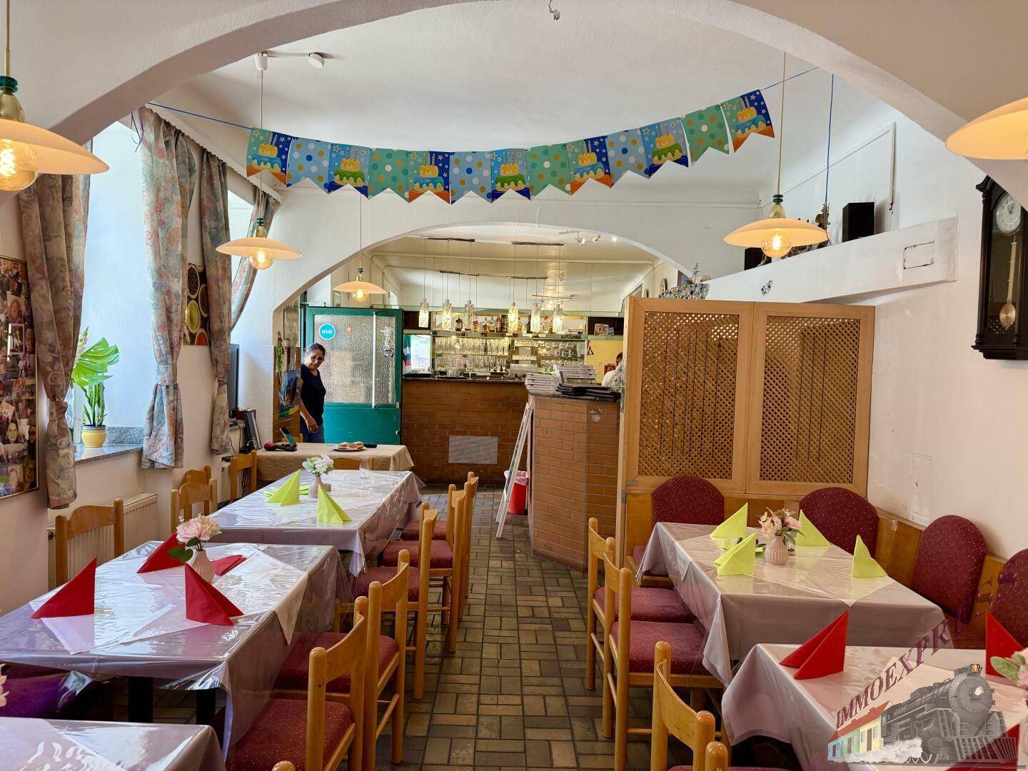 Langer Restaurant-Speisesaal mit mehreren Tischen, Stühlen und einer Bar im hinteren Bereich, geschmückt mit Partygirlanden.