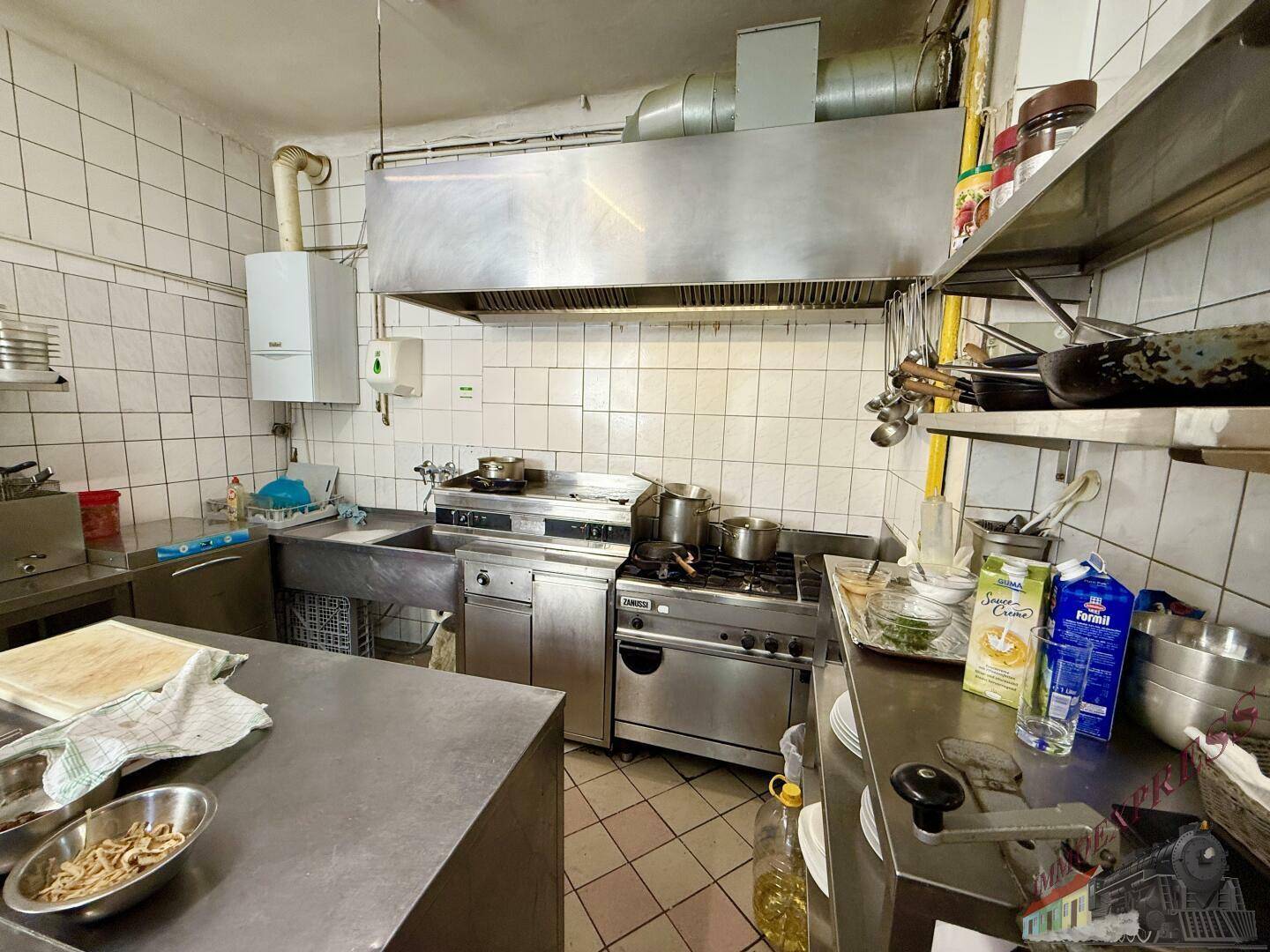 Kommerzielle Restaurantküche mit Edelstahl-Arbeitsflächen, Gasherd und einer großen Dunstabzugshaube über dem Kochbereich.