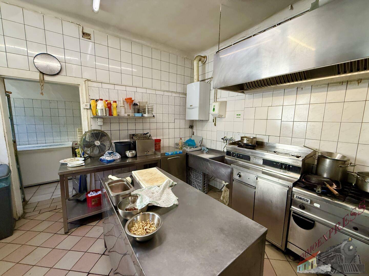 Kommerzielle Restaurantküche mit Edelstahl-Arbeitsflächen, Gasherd und einer großen Dunstabzugshaube über dem Kochbereich.