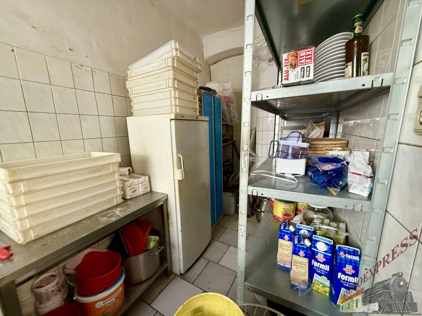Lagerbereich einer Restaurantküche mit Regalen, Kühlschrank und gestapelten Pizzakartons und Vorräten.