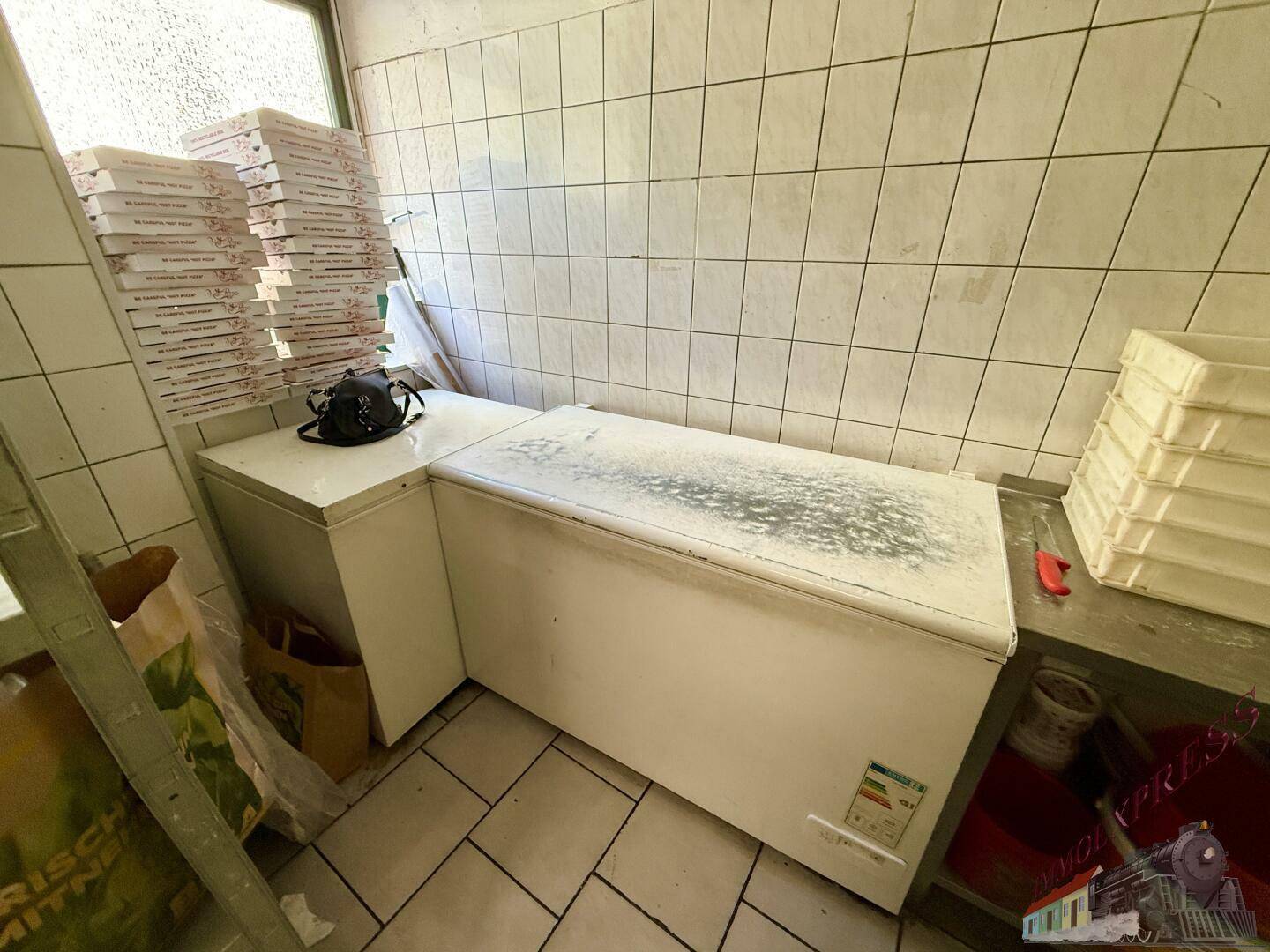 Lagerbereich einer Restaurantküche mit großen Gefriertruhen und gestapelten Pizzakartons neben einem Fenster.