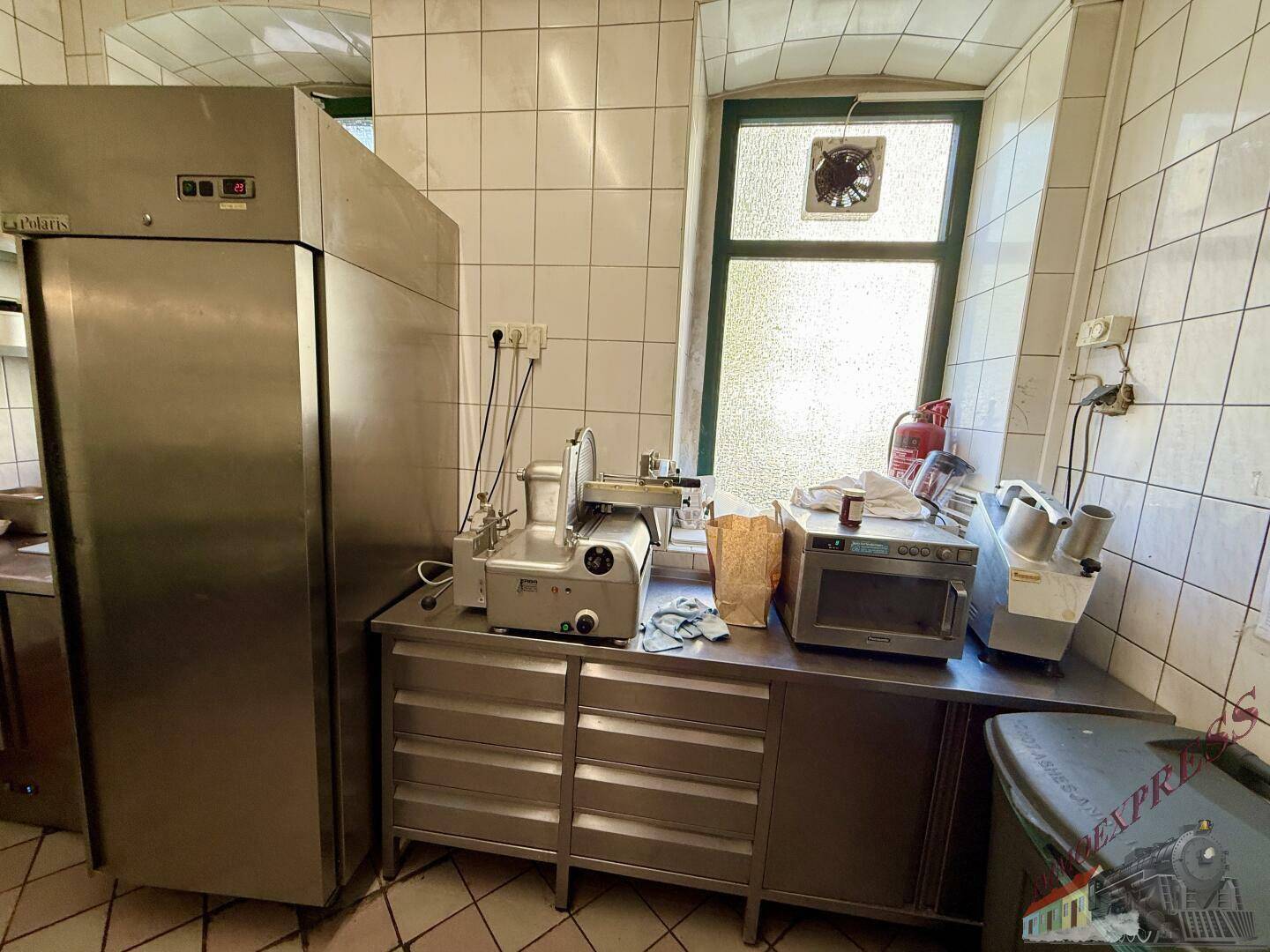 Professionelle Restaurantküche mit Edelstahlgeräten, Fliesenwänden und einem Fenster mit Abluftventilator.