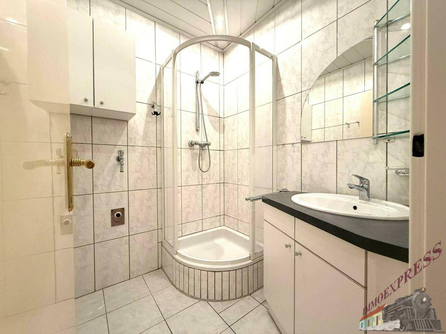 Badezimmer mit Duschkabine, Waschbecken und hellen Fliesen, praktisch und gut erhalten.