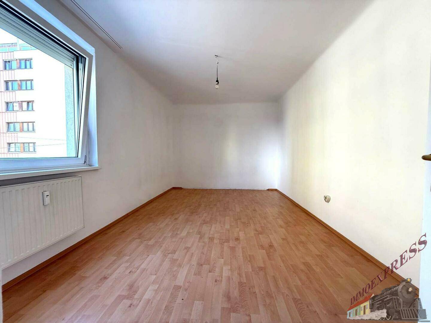Helles Zimmer mit Parkettboden und großem Fenster, das viel Tageslicht bietet.