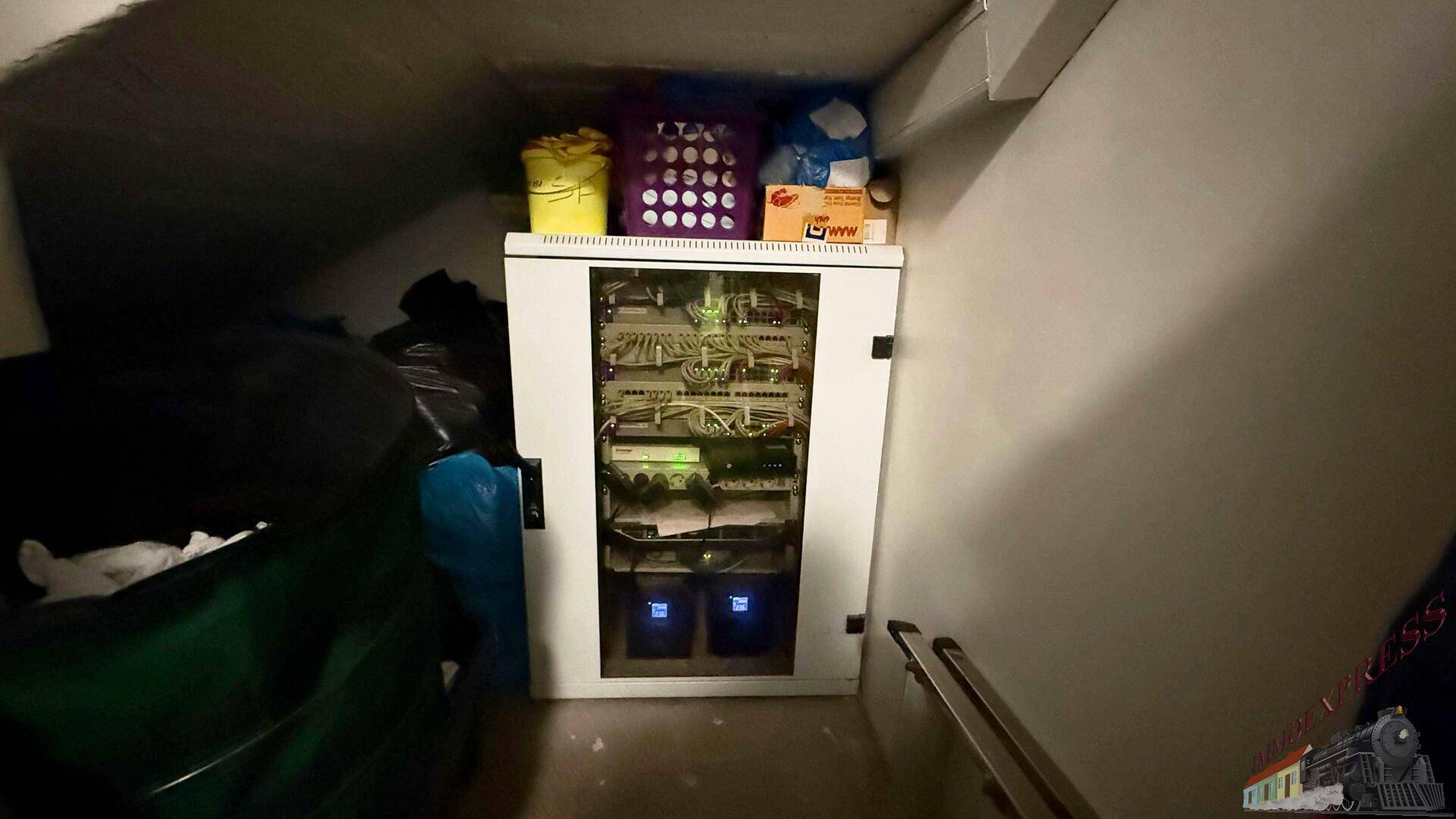 Kleiner Abstellraum mit einem Serverschrank und verschiedenen gelagerten Gegenständen unter einer Treppe.