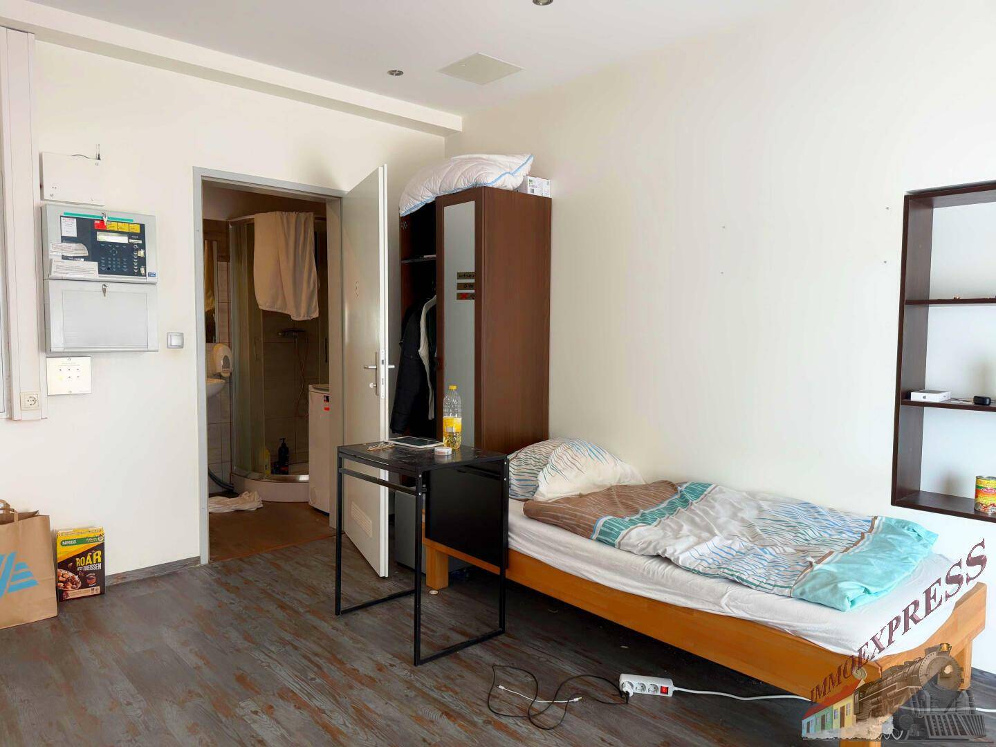 Einfaches Schlafzimmer mit Einzelbett, Kleiderschrank und Holzboden, funktional und hell gestaltet.