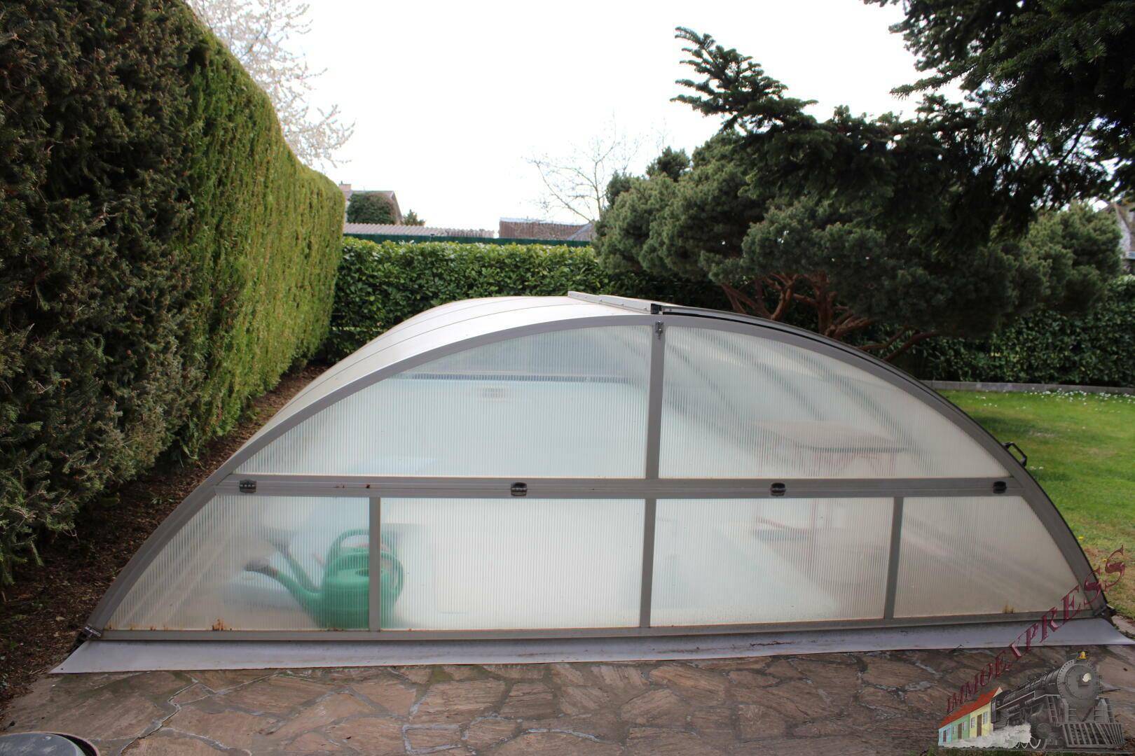 Überdachter Swimmingpool im Gartenbereich, umgeben von Hecken und Grünflächen.