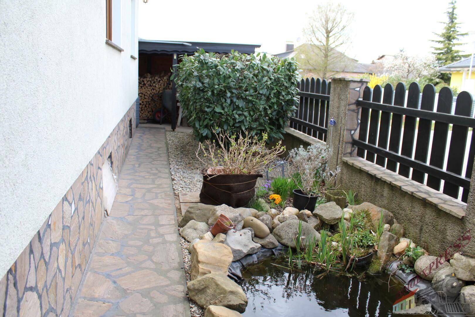 Seitlicher Gartenweg mit einem kleinen Teich und Holzstapel im Hintergrund.