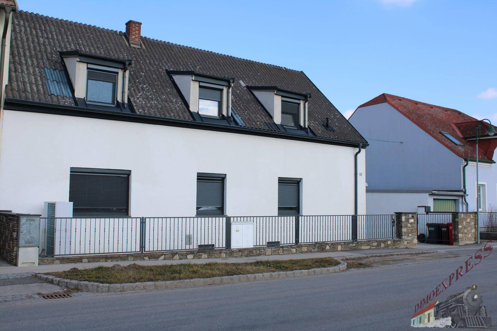 Vorderansicht des weißen Hauses mit Satteldach, Dachgauben und Steinmauer.