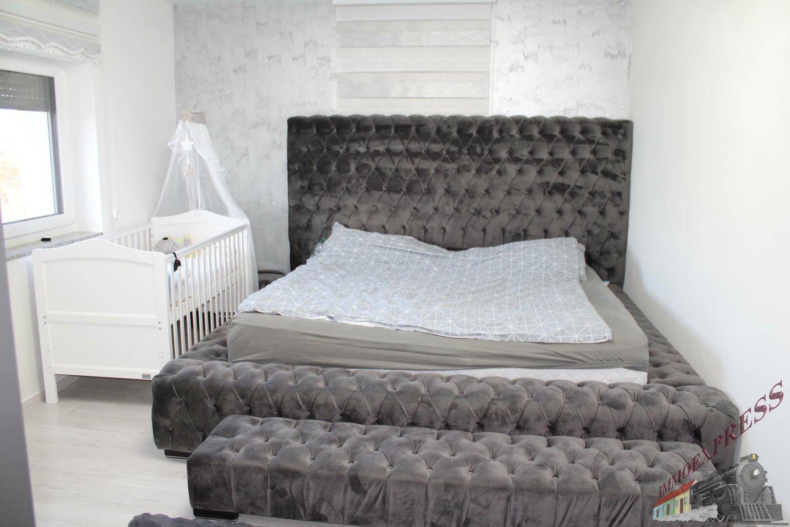 Schlafzimmer mit großem, gepolstertem Bett in Grau und einem Babybett am Fenster.