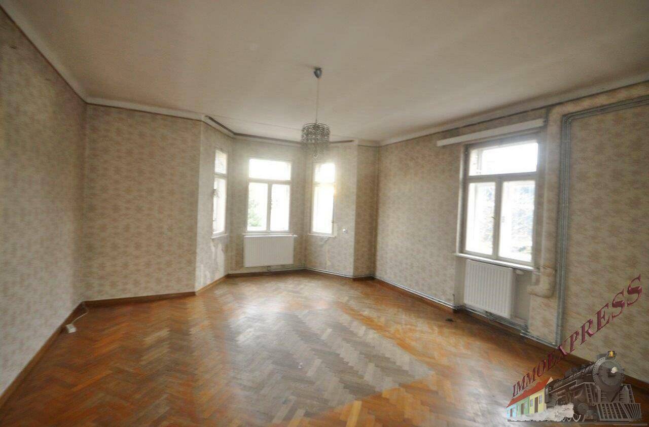 Geräumiges Wohnzimmer mit Fischgrätparkett, gemusterter Tapete und einem Erkerfenster mit Kronleuchter.