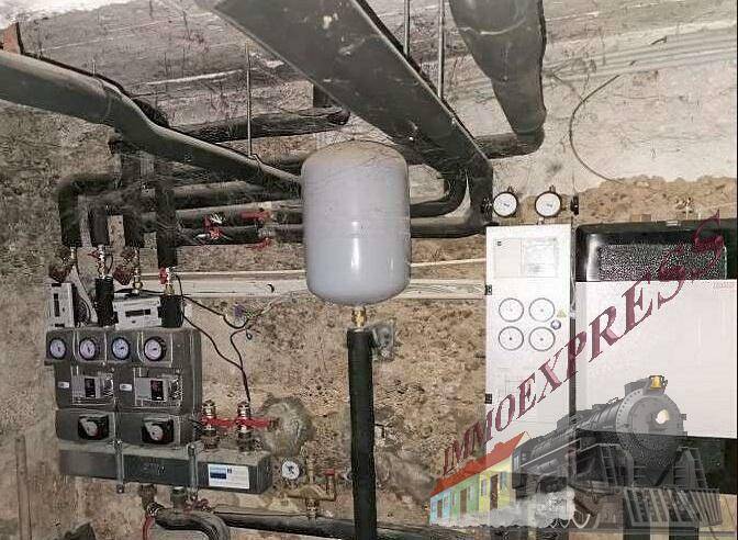 Technikraum im Keller mit sichtbaren Rohren, einem Warmwasserspeicher und verschiedenen Steuerungseinheiten.