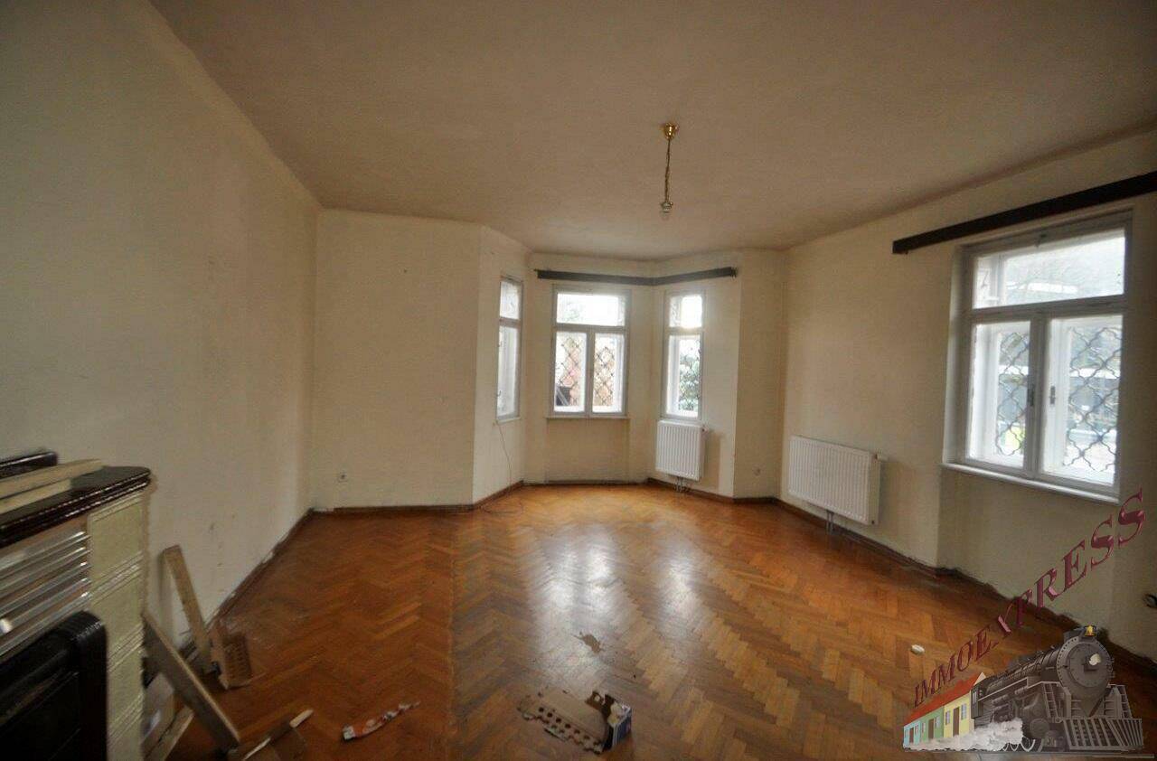 Geräumiges Wohnzimmer mit Fischgrätparkett, Erkerfenstern und einem traditionellen Ofen.