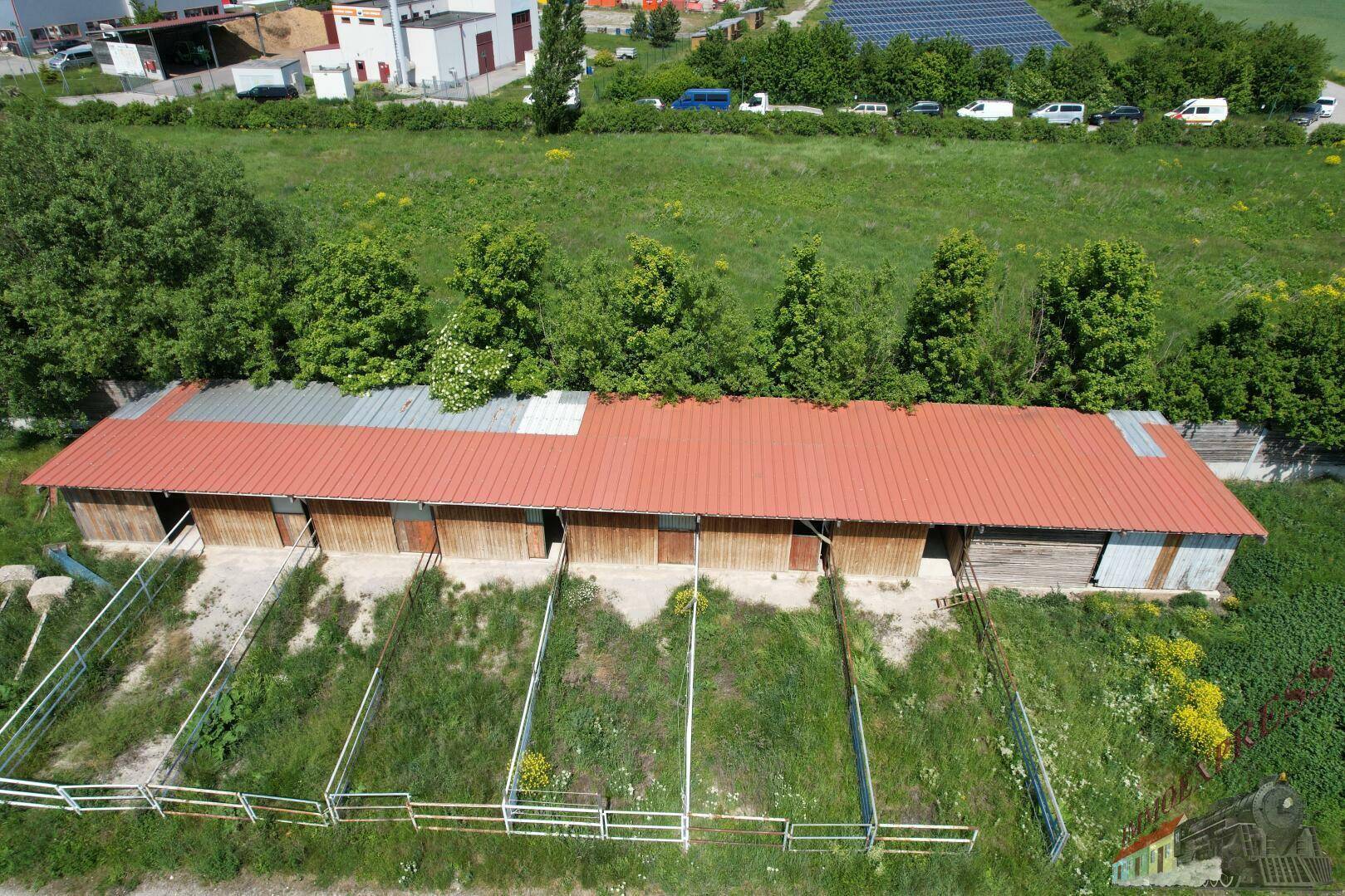 Luftaufnahme eines langen Gebäudes mit roten Dächern und mehreren Boxen oder Ställen, umgeben von Grünflächen.