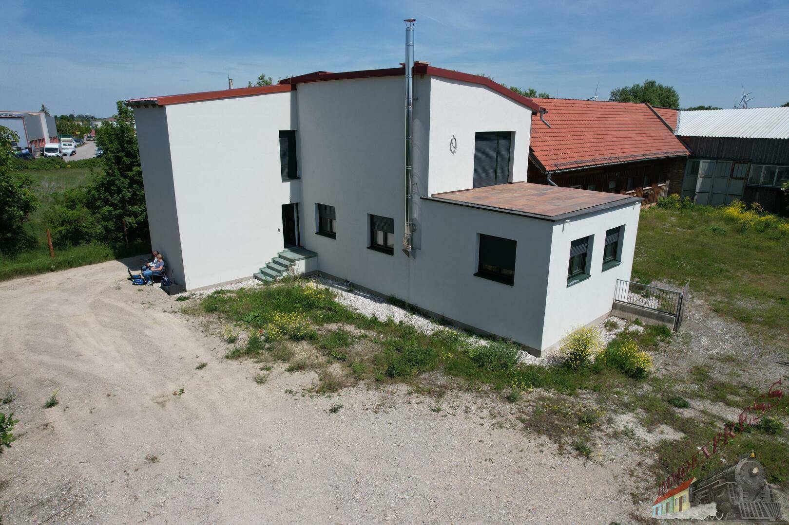 Ansicht eines modernisierten weißen Gebäudes mit Flachdach und kleinen Fenstern, umgeben von einer Schotterfläche.