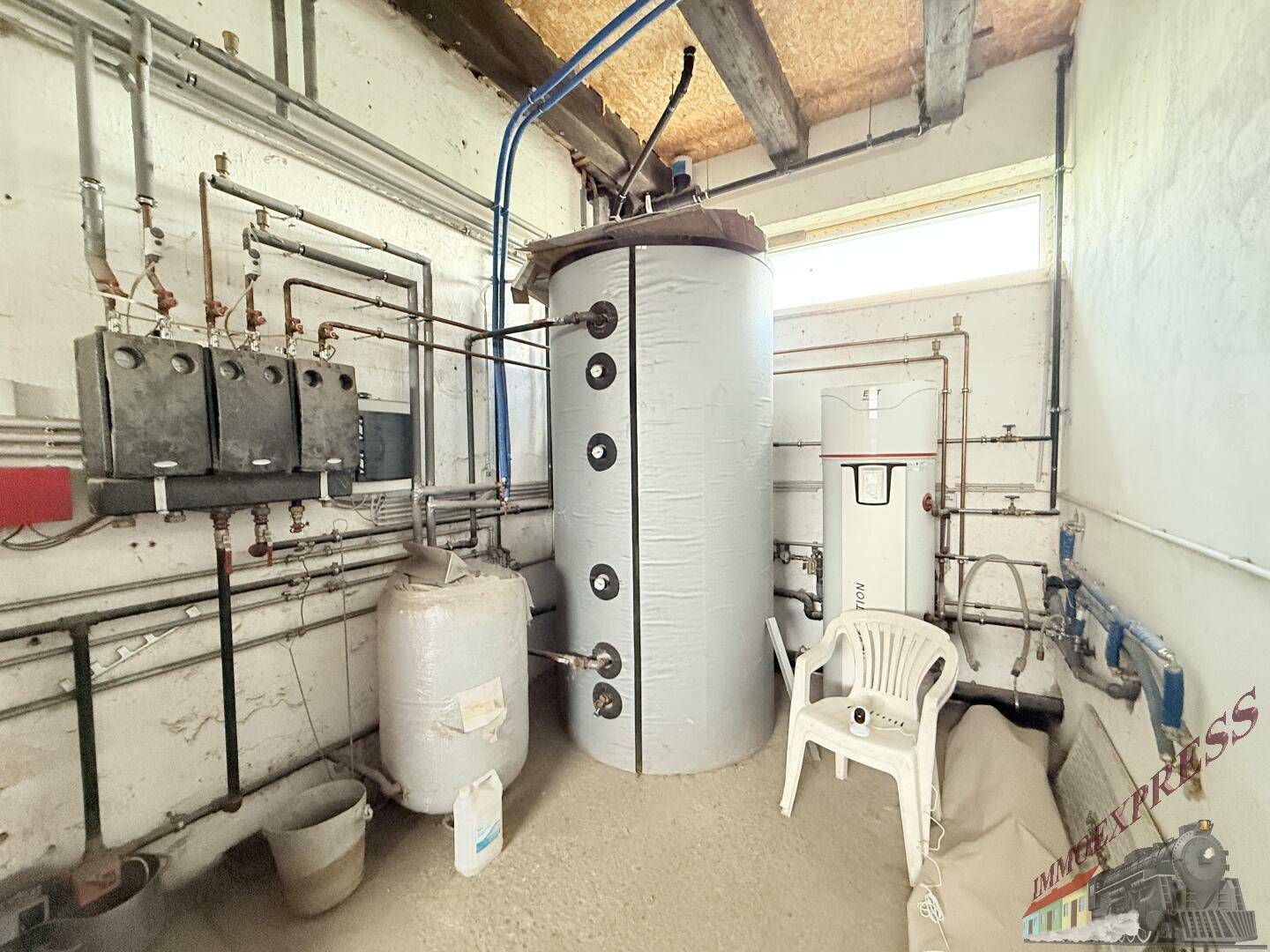 Technikraum mit großem Warmwasserspeicher und komplexer Rohrleitungsinstallation, unter Holzbalkendecke.