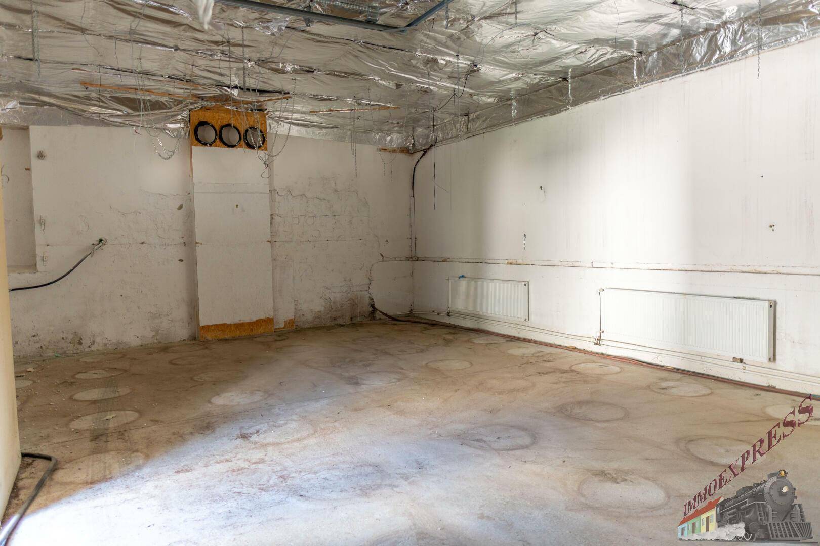 Unfertiger Kellerraum mit Betonboden, weißen Wänden und sichtbaren Installationen an der Decke.