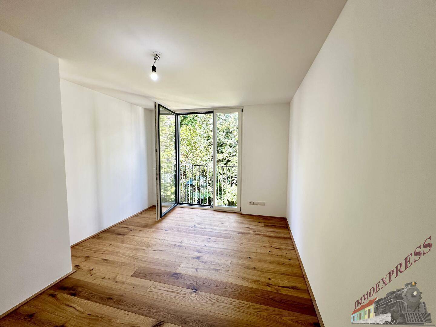 Helles Zimmer mit Holzboden und Zugang zu einem französischen Balkon mit Grünblick.