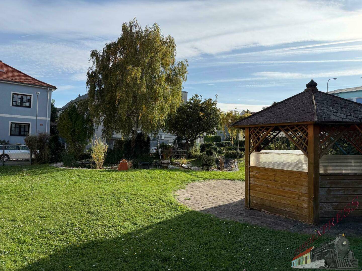 Sonniger Garten mit einer hölzernen Laube und weitläufiger Rasenfläche.