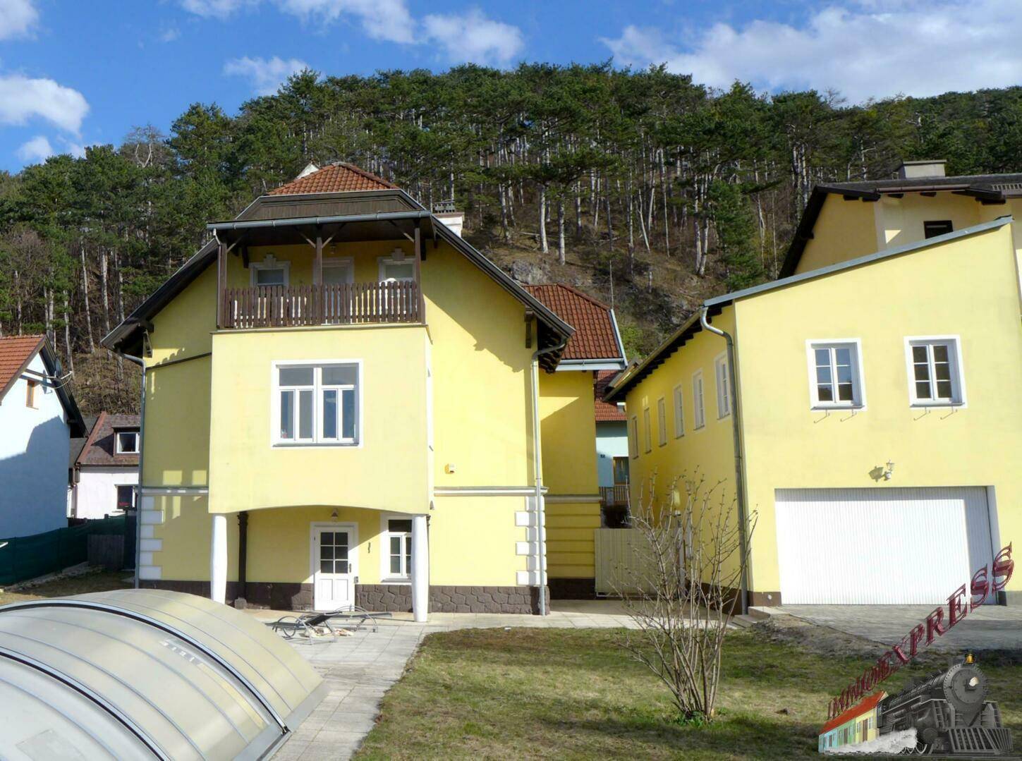 Gelbes Wohnhaus mit Balkon, Garage und Gartenbereich vor bewaldetem Hügel.