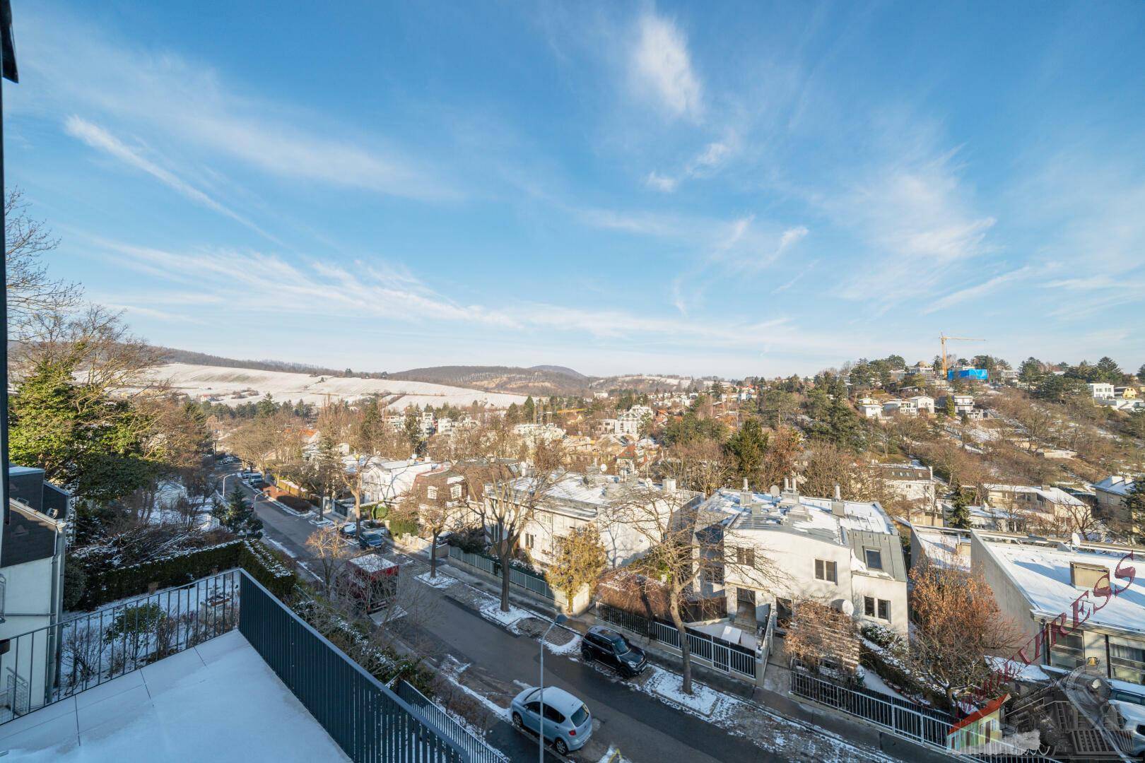 Panoramablick über eine verschneite Wohngegend mit Häusern und grünen Hügeln unter blauem Himmel.