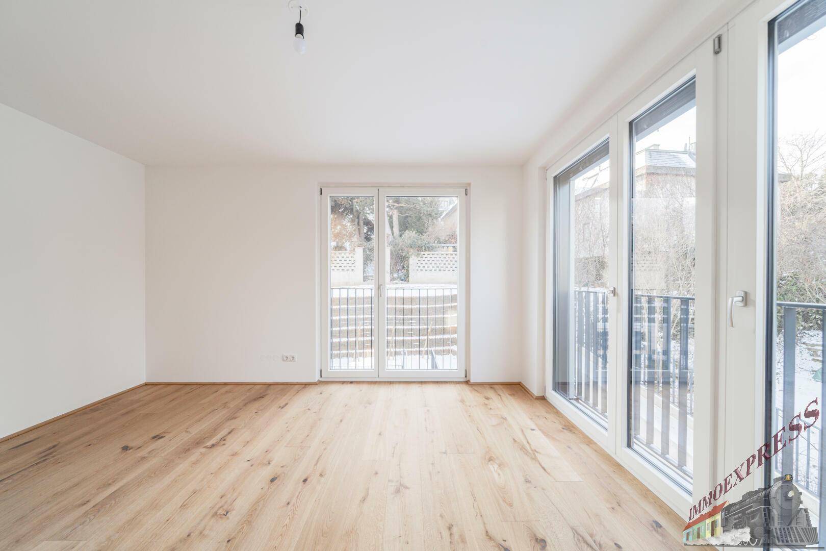 Helles Zimmer mit Holzboden und großen Fenstern, die viel Tageslicht hereinlassen und einen grünen Ausblick bieten.