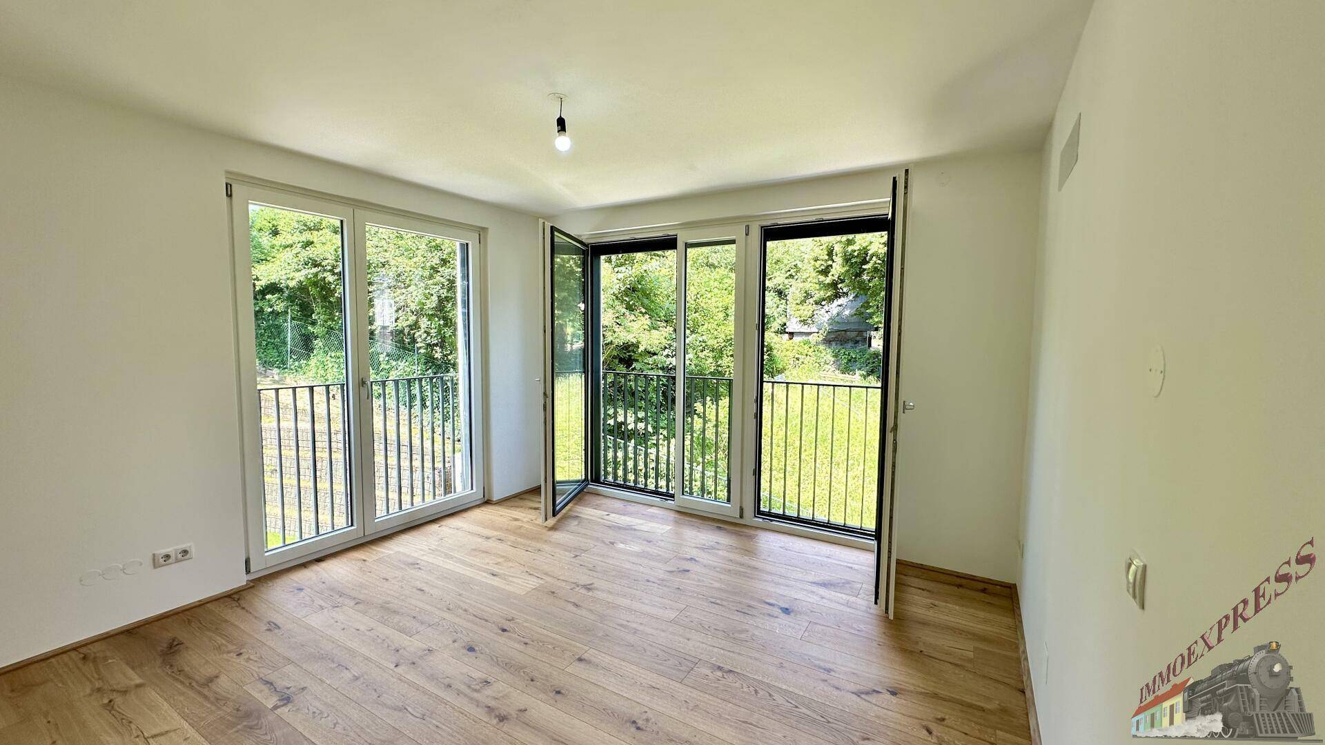 Helles Zimmer mit Holzboden und großen Fenstern, die viel Tageslicht hereinlassen und einen grünen Ausblick bieten.