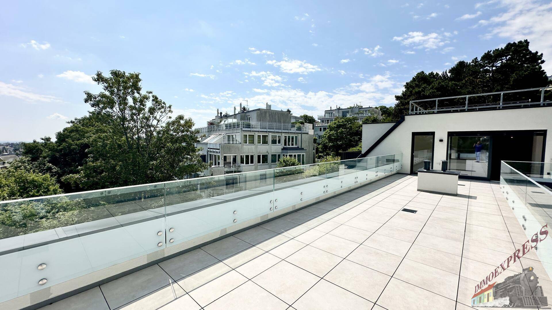 Sonniger Terrassenbereich mit Glasgeländer und Blick auf die umliegenden Gebäude und Bäume.
