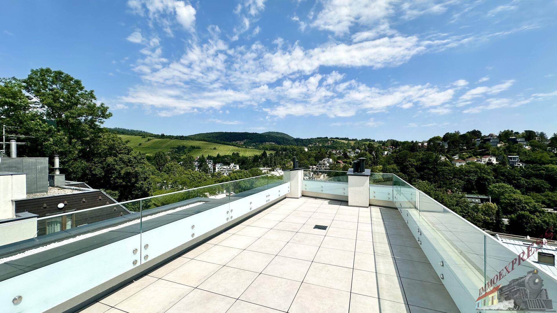 Große Dachterrasse mit Fliesenboden und Glasgeländer, umgeben von grüner Hügellandschaft.