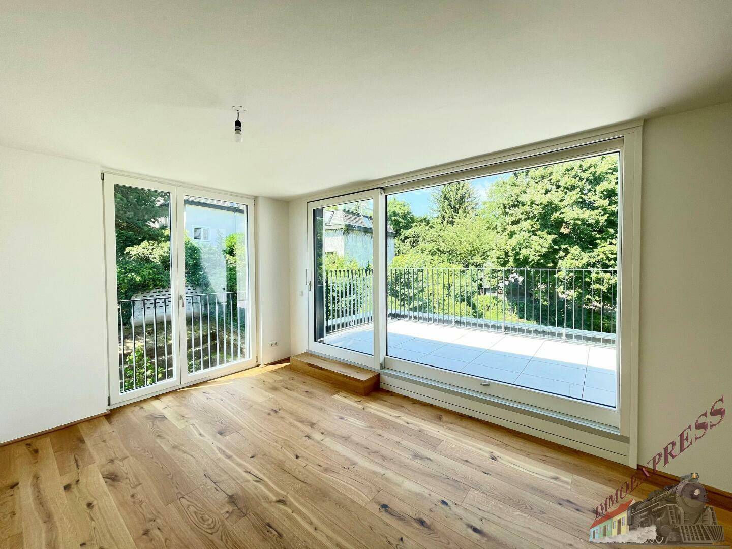 Helles Zimmer mit Holzboden und großen Fenstern, die auf eine grüne Terrasse führen.