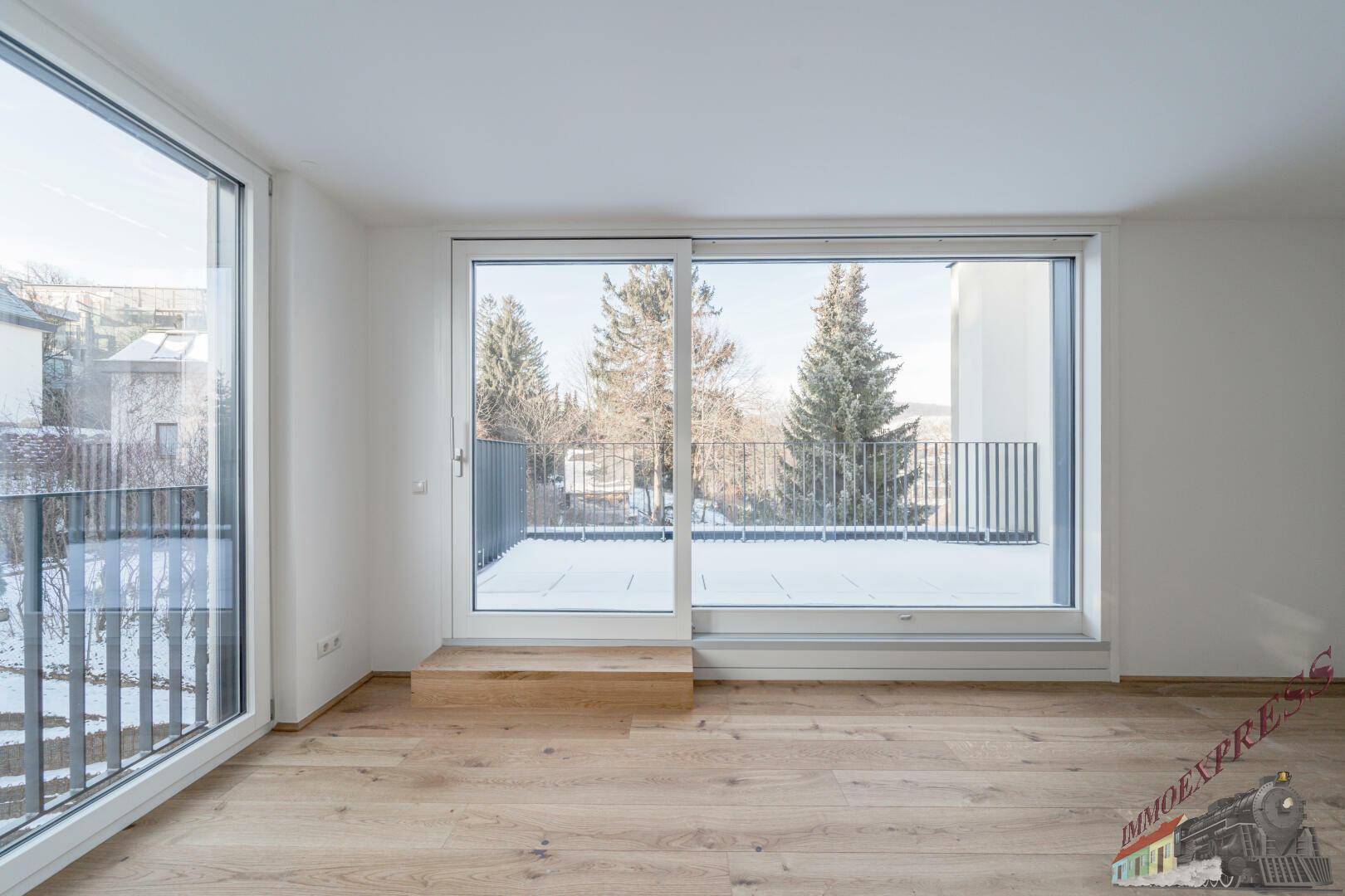 Heller Raum mit Holzboden und großen Fenstern, die einen Blick auf die Umgebung bieten.