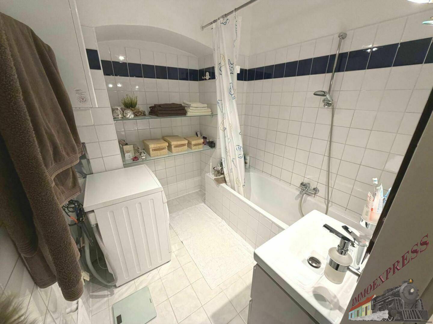 Gefliestes Badezimmer mit Badewanne, Dusche und Waschmaschine, ausgestattet mit praktischen Ablagen.