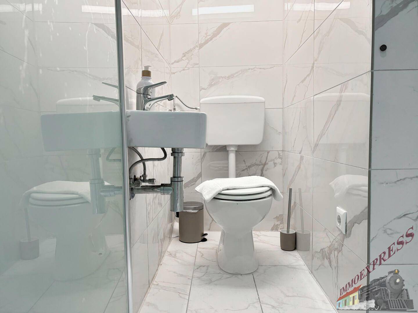 Kompaktes Badezimmer mit WC, kleinem Waschbecken und durchgehenden weiß-marmorierten Fliesen.