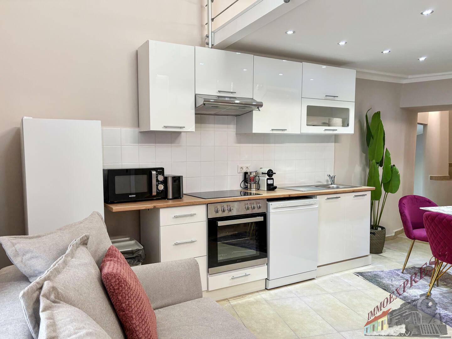 Kompakte Küche mit weißen Schränken und integrierten Geräten, offen zum Wohnbereich mit Sofa.