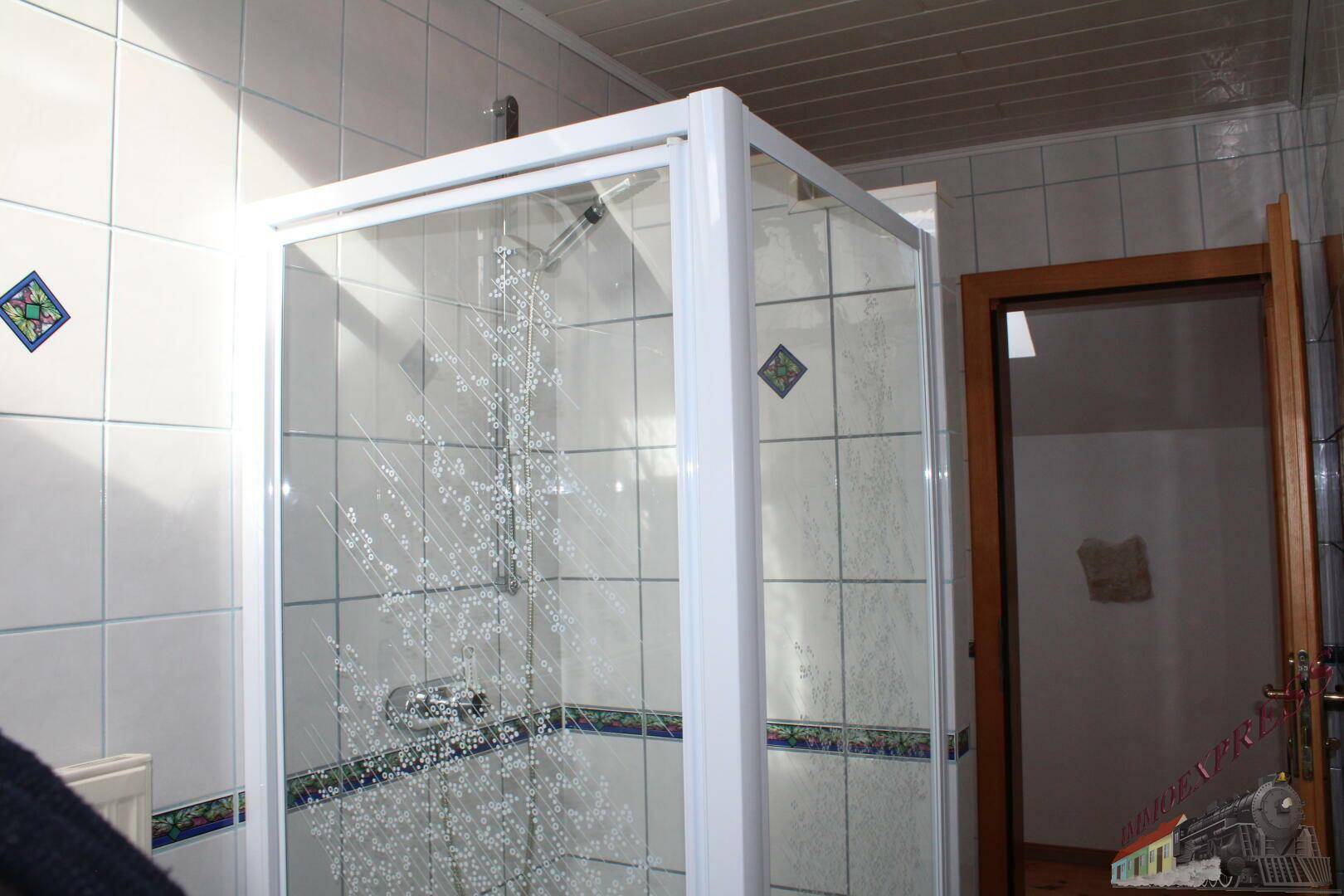 Badezimmer mit Duschkabine und weißen Fliesen, teilweise mit blauen Akzenten und gemusterten Bordüren.