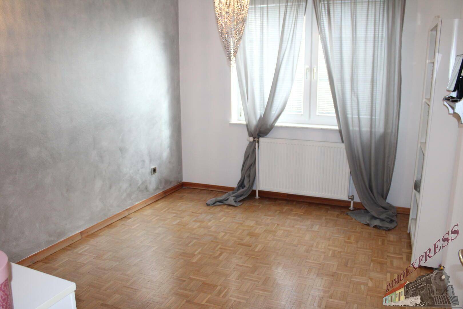 Leeres Zimmer mit Parkettboden, grauer Akzentwand und großem Fenster für viel Tageslicht.