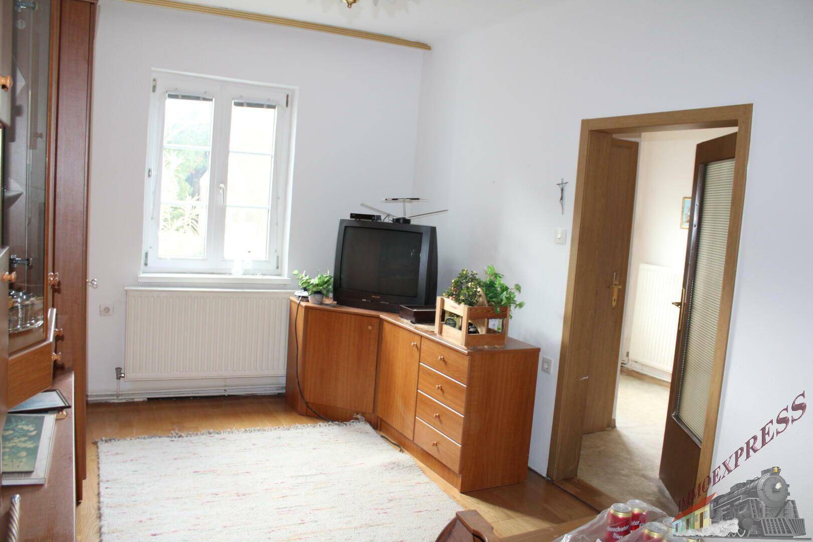 Wohnzimmer mit Holzschrank, Fernseher und Fenster mit Blick ins Grüne, hell und freundlich.