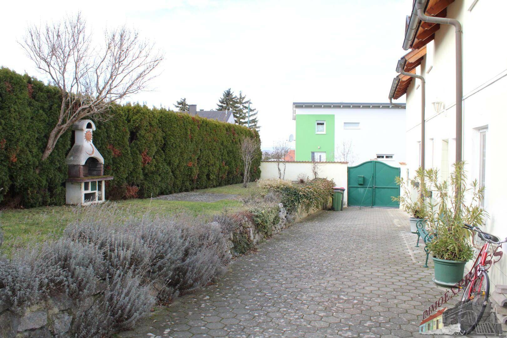 Gartenbereich mit gepflasterter Zufahrt, hoher Hecke und gemauertem Grillplatz.